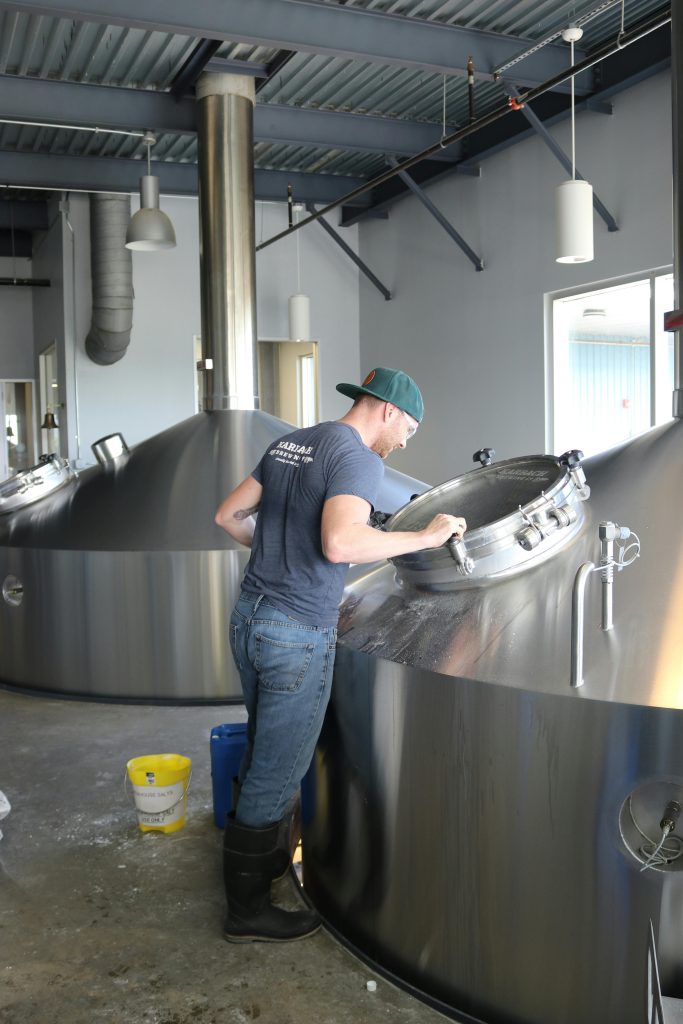A worker in a brewery inspects large stainless steel fermentation tanks.